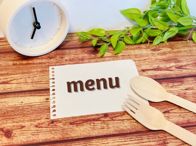 木目調のテーブルの上に置かれた「menu」と書かれた紙、白い置時計、木製のフォークとスプーン。メニュー構成や時間帯による客単価向上の仕組みを示唆する画像。