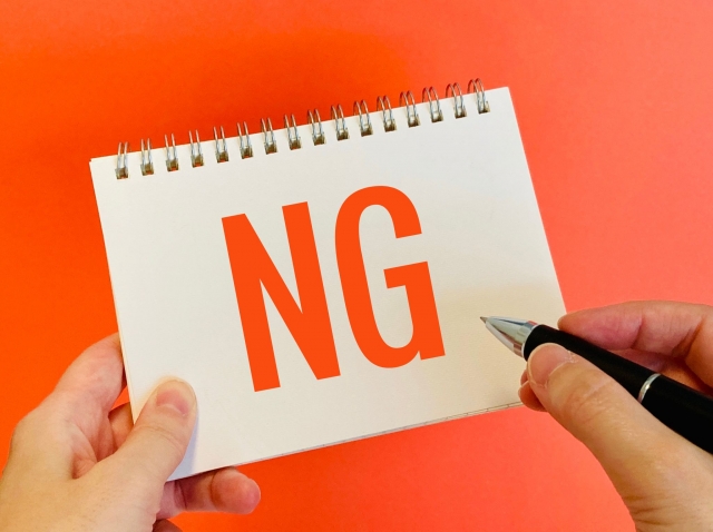 ノートに大きく「NG」と書かれたメモを持つ手の写真。やってはいけない行動やNGワードを示すイメージ。