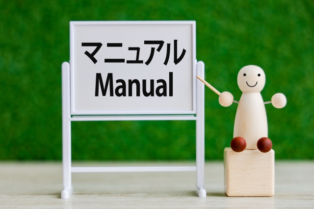 マニュアルを指し示す木製のキャラクターの写真。仕事の基本やルールの重要性を表すイメージ。