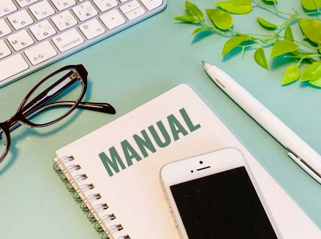 ノートに「MANUAL」と書かれた冊子とスマートフォン、眼鏡、ペンが並ぶデスクの写真。マニュアル作成や教育をイメージした構図。