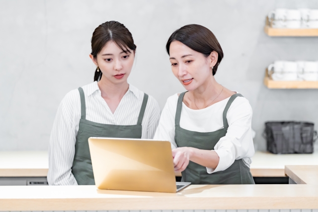 カフェのカウンターでスタッフがパソコンを見ながら仕事の内容を確認している様子。業務マニュアルの共有をイメージした写真。